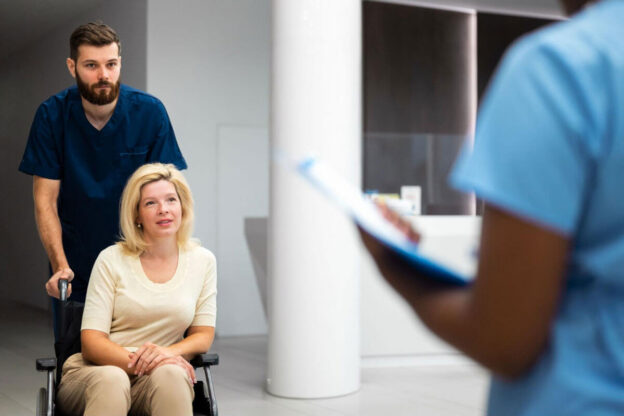 A male nurse in dark scrubs pushes a woman in a wheelchair through a bright clinical hallway toward another healthcare worker holding a clipboard.